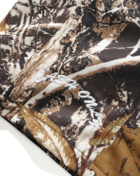 Close-up of a camouflage-patterned garment, likely shorts, featuring a brown, beige, and black woodland camo print with white embroidered script text on the side.