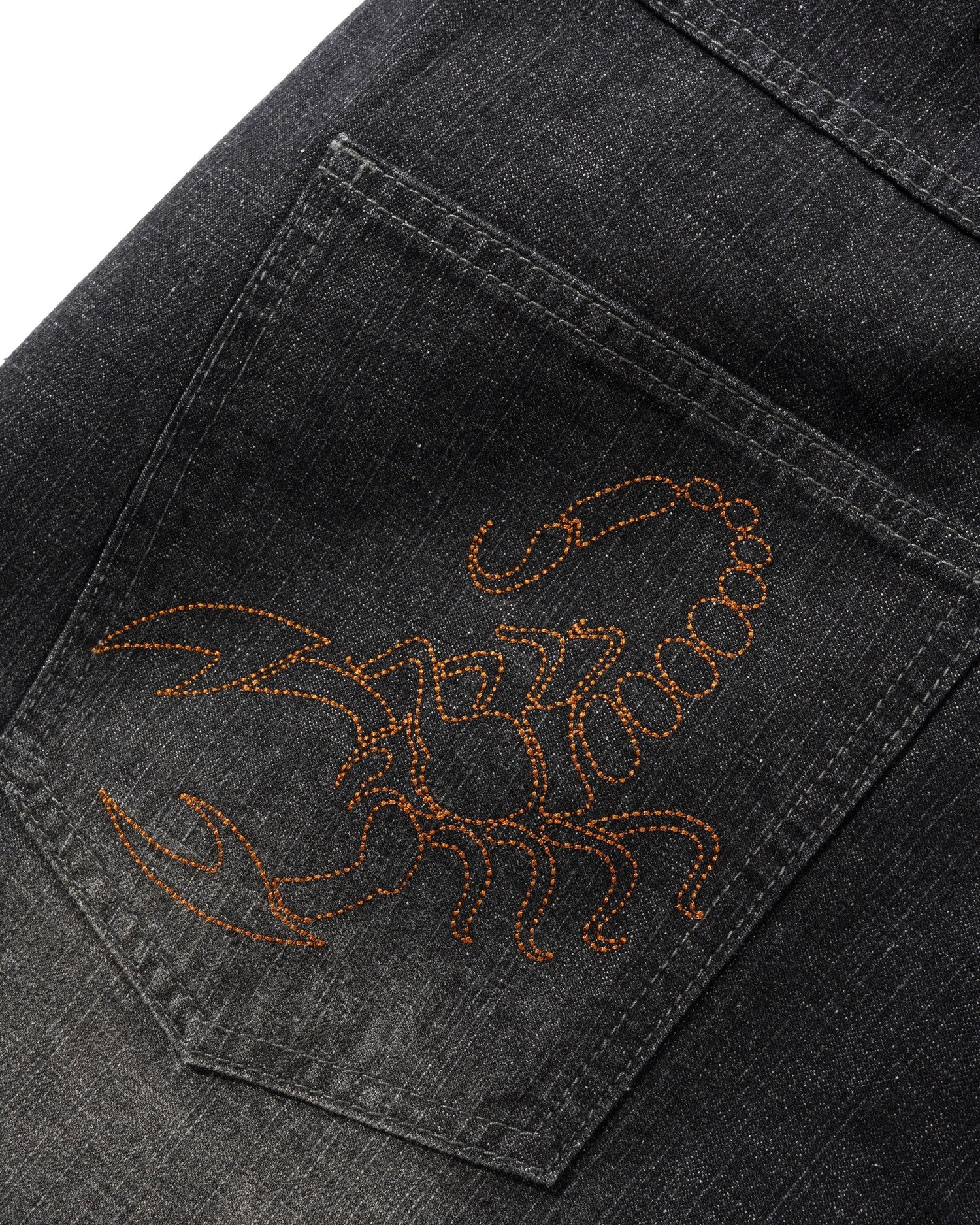 Close-up of a dark gray denim jeans pocket featuring an orange-brown embroidered scorpion design on the back.