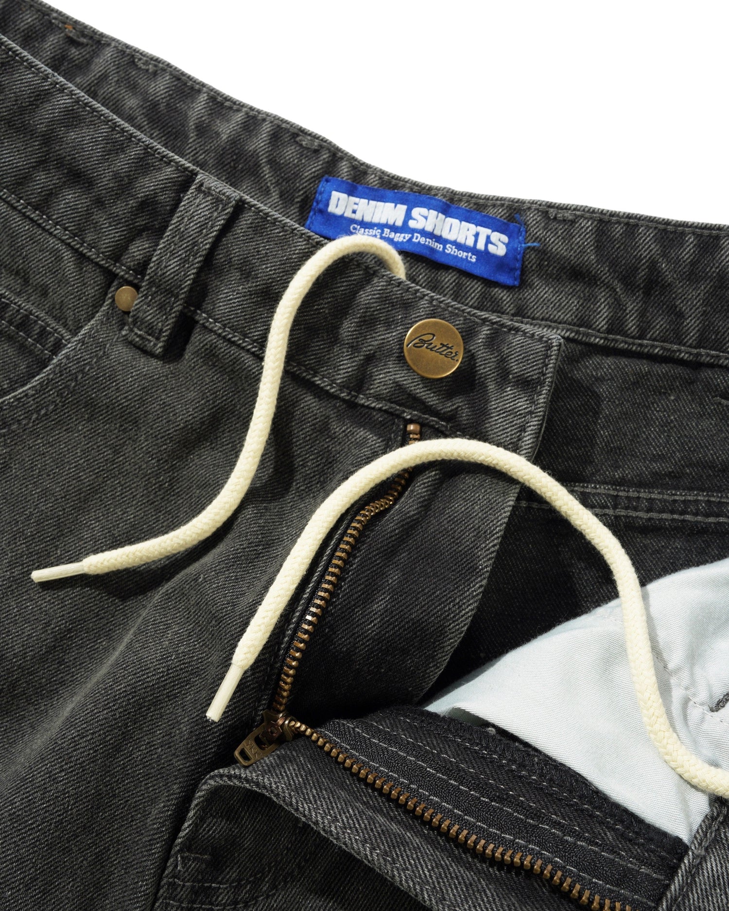 Close-up of gray denim shorts with brand label and zipper detail.